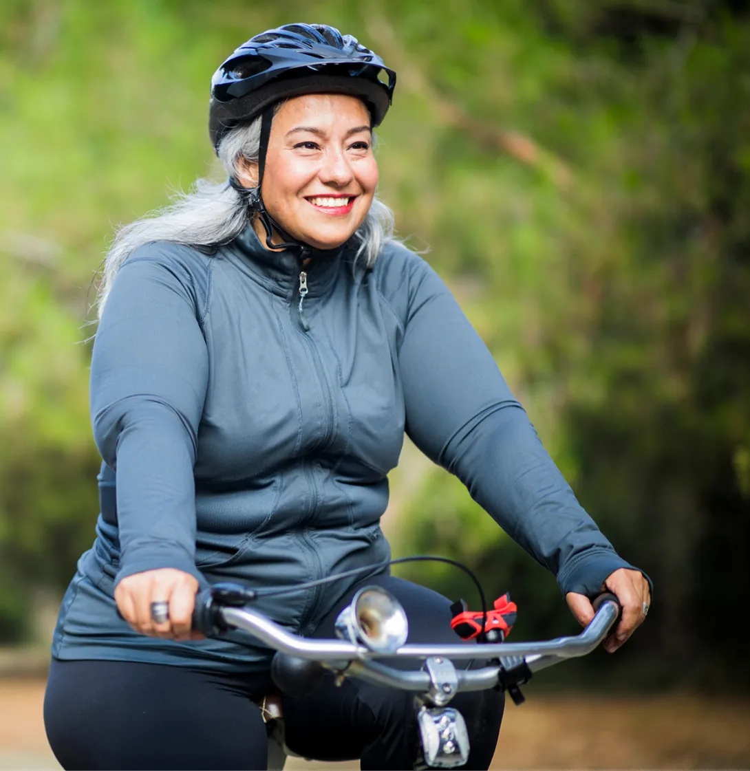 An adult smiling while cycling outdoors on a tree-lined trail.